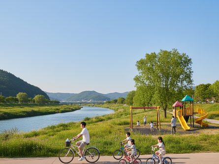 맑은 하늘 아래 푸른 강변 산책로, 자전거 타는 가족과 놀이터, 측면 촬영